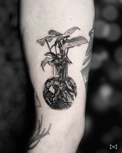 Planta com raízes aparentes dentro de um vaso de vidro, tatuada no braço
